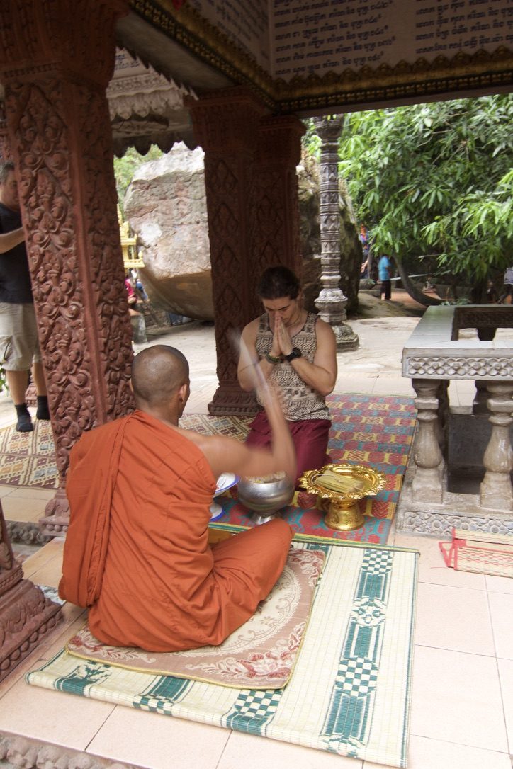 A volunteer receives a blessing from a monk in an orange robe at a carved stone temple in Cambodia